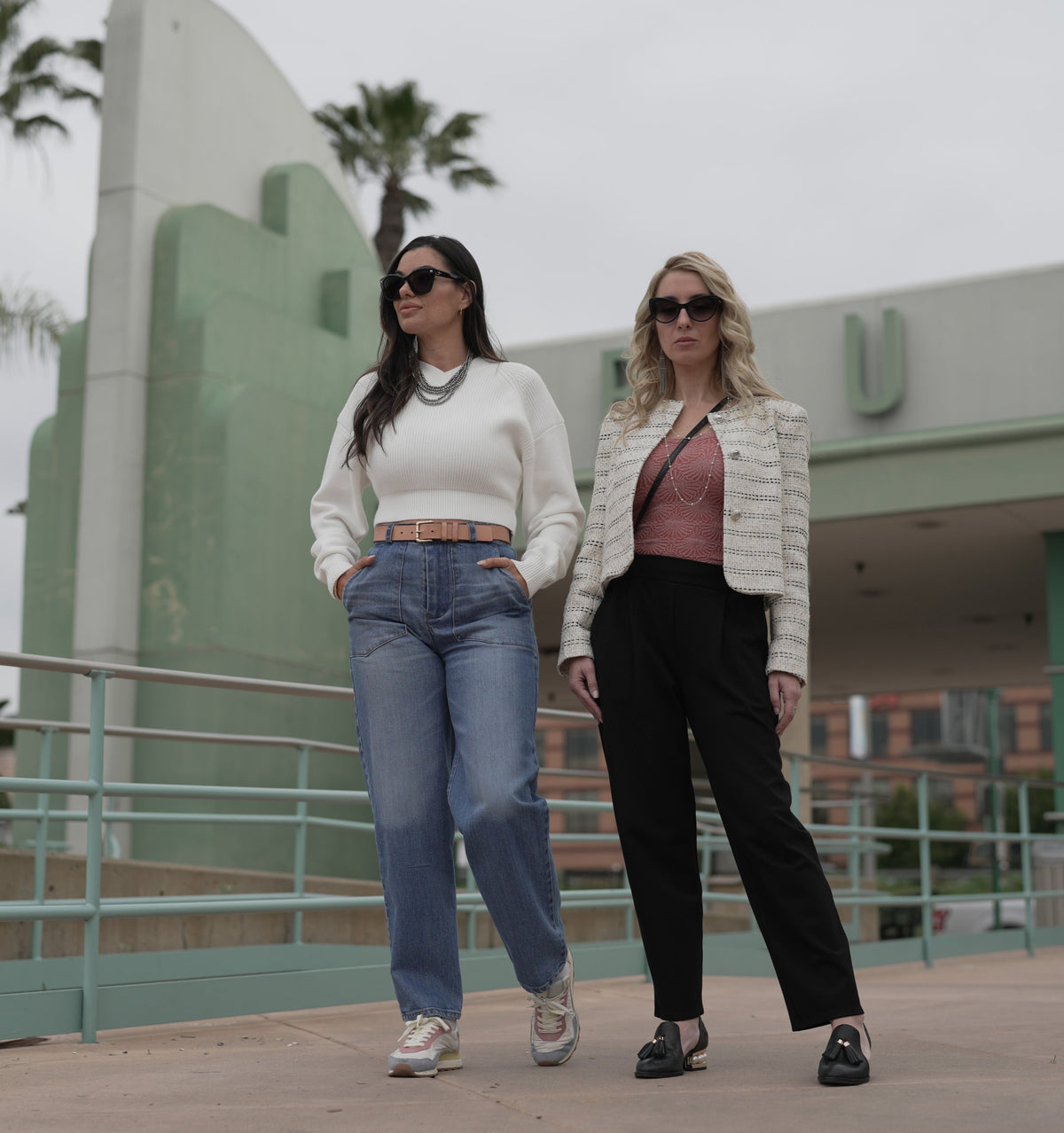 murjer women posing at a train station. one wears Austin, a women's barrel leg denim pant, high rise, fitted waist and hips with a relaxed barrel leg and dart knee detail. medium blue color and wash. functional zipper and button. the other woman wears the brooklyn women's ponte knit tapered pull-on pant with high rise, fitted elastic waist in black. this has a looser fit in the hips with front pleats and pockets. Includes built in shapewear to  define your waist and enhance and lift your booty.