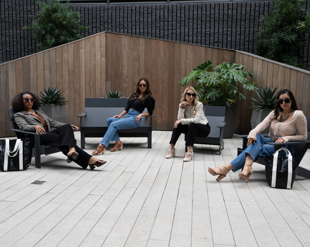 four stylish ladies sitting in architectural courtyard wearing MURJER slimming, sculpting fashion pants and showcasing murjer reusable tote bags. pants featured are morrison wide leg in midnight, venice straight leg denim pant in blue quartz, brooklyn tapered ponte pant in obsidian, austin barrel leg in blue quartz. Murjer girls are impeccably accessorized in fashion heels, jewelry and sunglasses.