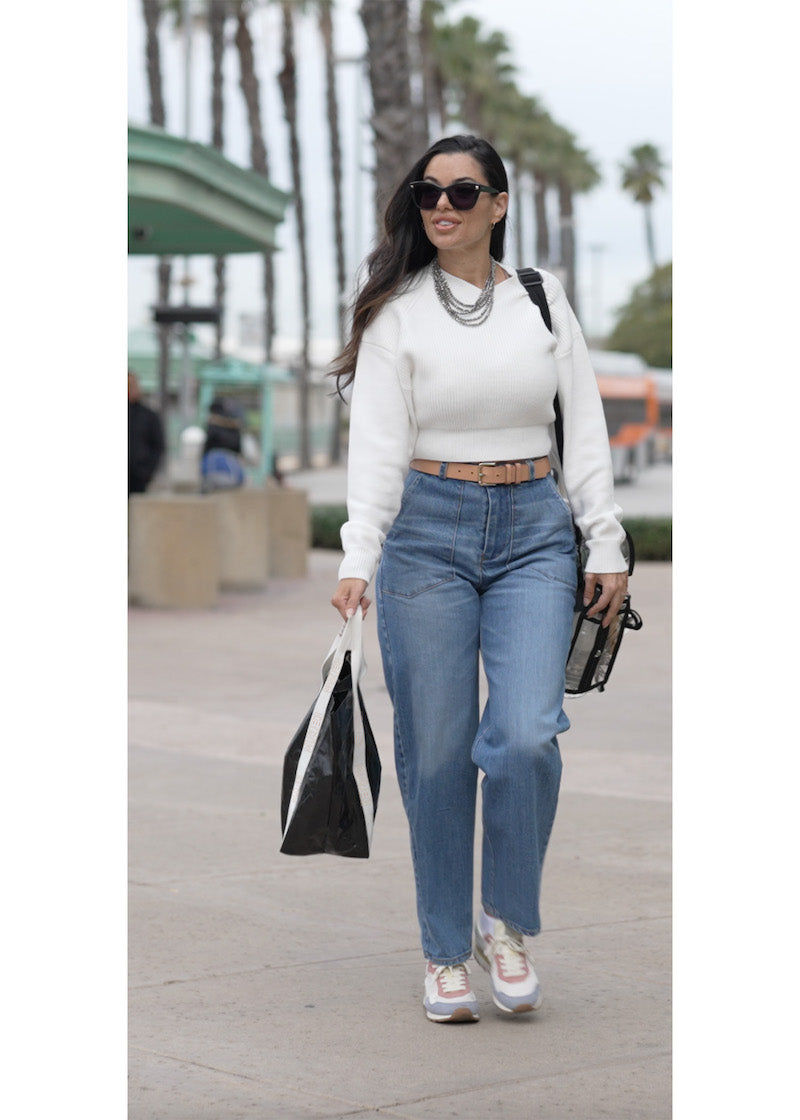 fashionable murjer make-up artist walking through a train station lined with palm trees. murjer woman is wearing the austin barrel leg jean in blue quartz styled with a white cropped sweater and accessorized with a silver layered necklace chain, tan colored belt, sporty sneakers and sunglasses provided by image optics. She is carrying a make-up bag and murjer reusable tote.